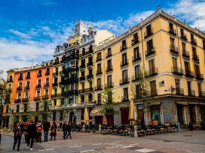 Edificios coloridos con balcones y toldos. La gente camina por la acera frente a ellos bajo un cielo azul.