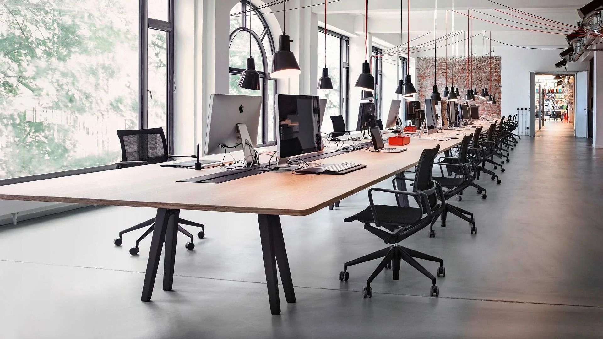 Mesa larga de madera con ordenadores y sillas negras en un espacio de oficina moderno, grandes ventanales y lámparas colgantes.