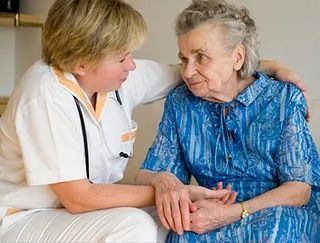 Una enfermera consuela a una anciana, sujetándole la mano y el brazo. Ambas están sentadas en el interior, vestidas con ropa profesional.