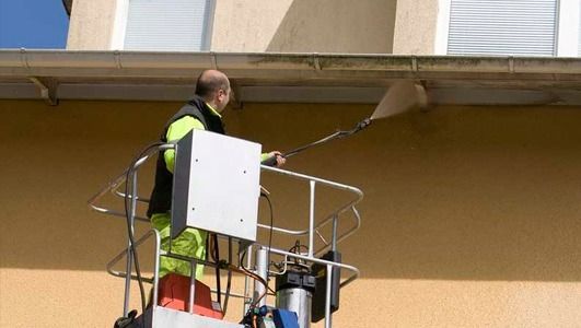 Hombre en una plataforma elevadora limpiando el canalón de un edificio con un cepillo de mango largo. Edificio amarillo, canalón marrón claro.