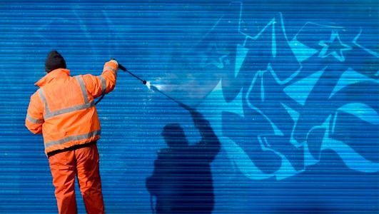Una persona vestida con un traje naranja limpia a presión un grafiti azul de una pared azul.