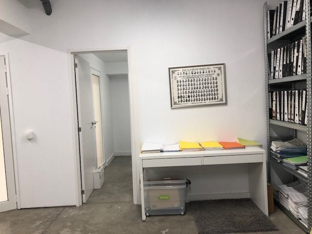 Una habitación blanca con un escritorio, un estante lleno de carpetas, un pequeño contenedor de almacenamiento y una puerta que conduce a otra habitación.