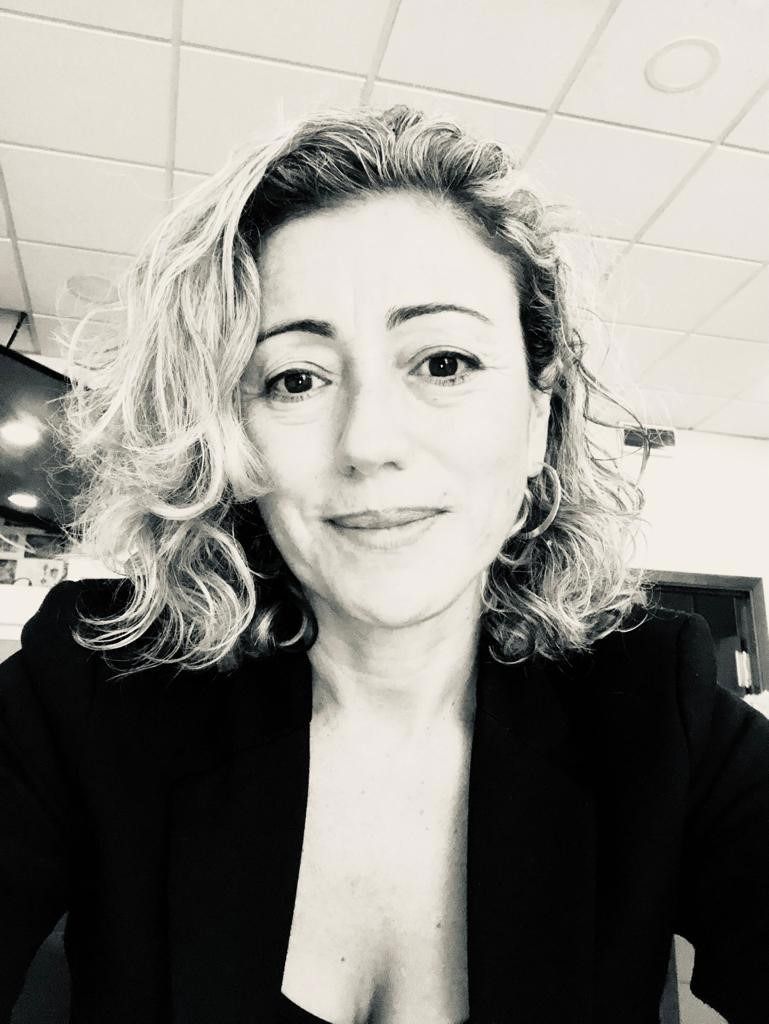 Una persona con cabello rizado hasta los hombros y vestida con una chaqueta oscura sonríe mientras posa para un retrato en un entorno de oficina.