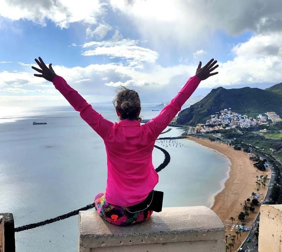 Una persona con una chaqueta rosa brillante está sentada en una cornisa con los brazos en alto, contemplando una playa, el océano y las montañas costeras.