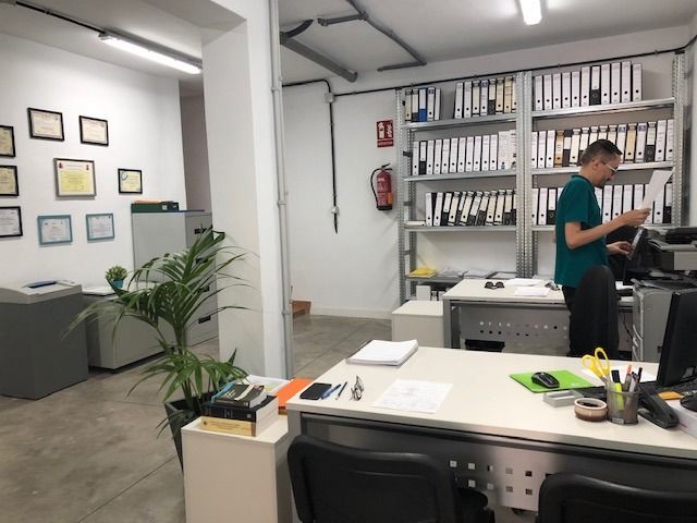 Escenario de oficina con una persona trabajando en un escritorio junto a estanterías llenas de carpetas y una pared decorada con certificados.
