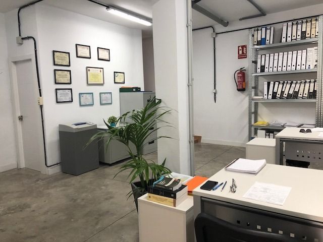 Una oficina con paredes blancas, certificados enmarcados, una planta, una estantería metálica con carpetas y escritorios en primer plano.