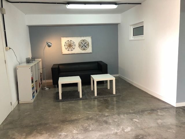 Una sala de estar con un sofá negro, dos mesas blancas, una estantería blanca y cuadros en una pared decorativa de color gris.