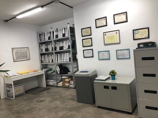 Una casa o una pequeña oficina con una estantería metálica llena de carpetas, un escritorio, una trituradora de papel y premios enmarcados en la pared.