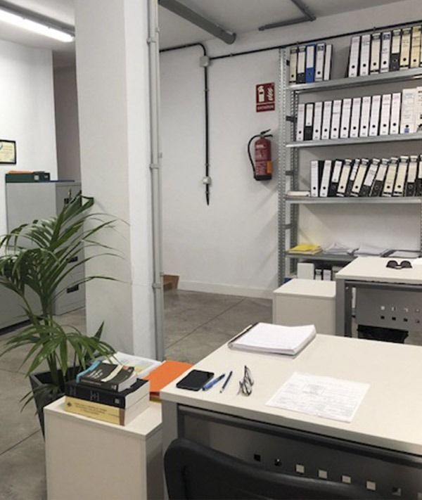 Un espacio de oficina ordenado con un escritorio con material de oficina, una planta de interior cerca y una estantería metálica llena de carpetas.