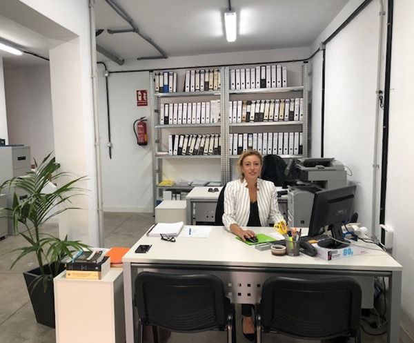 Una persona está sentada en un escritorio de oficina con una computadora, con estanterías llenas de carpetas y un extintor de incendios al fondo.