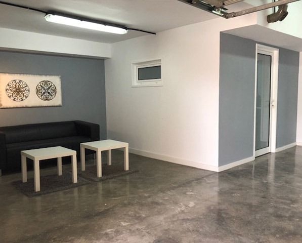 Una moderna sala de estar con un sofá negro, dos mesas blancas y obras de arte en las paredes, en una habitación con paredes grises y suelo de hormigón.