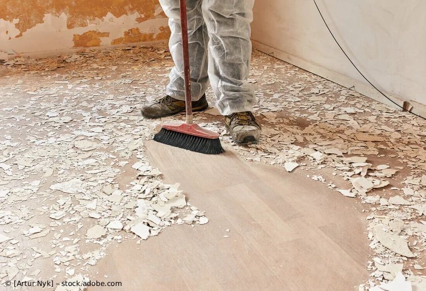 Eine Person in Schutzkleidung fegt Schutt von einem Holzboden in einem Zimmer.