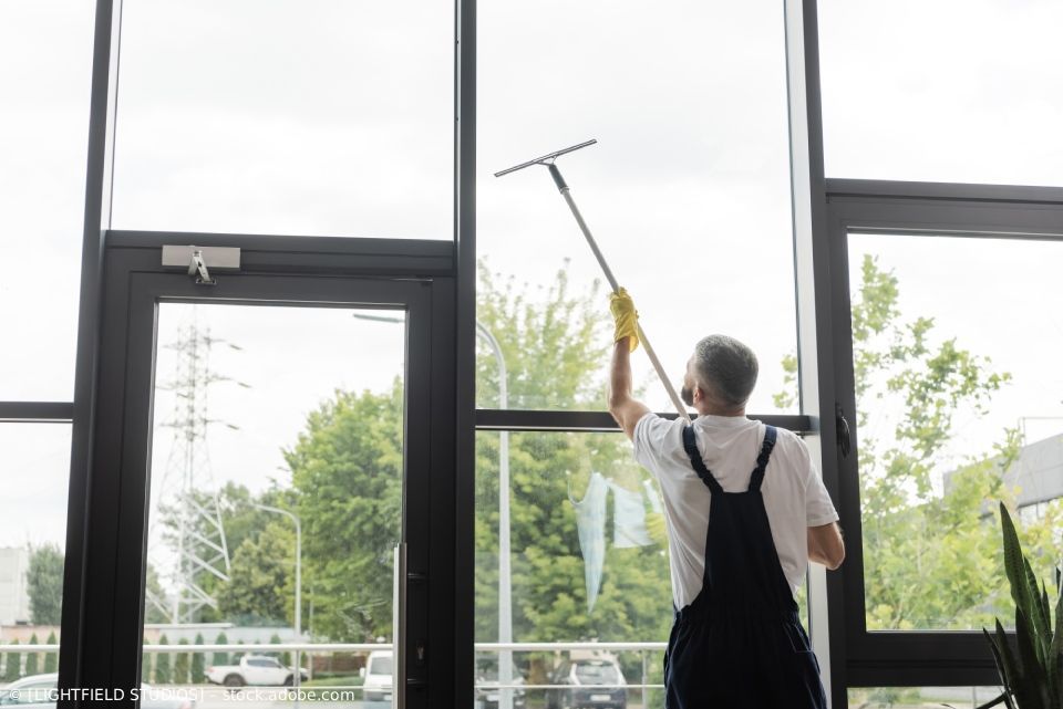Eine Person in Arbeitskleidung putzt mit einem Abzieher ein großes Fenster in einem Gebäude.
