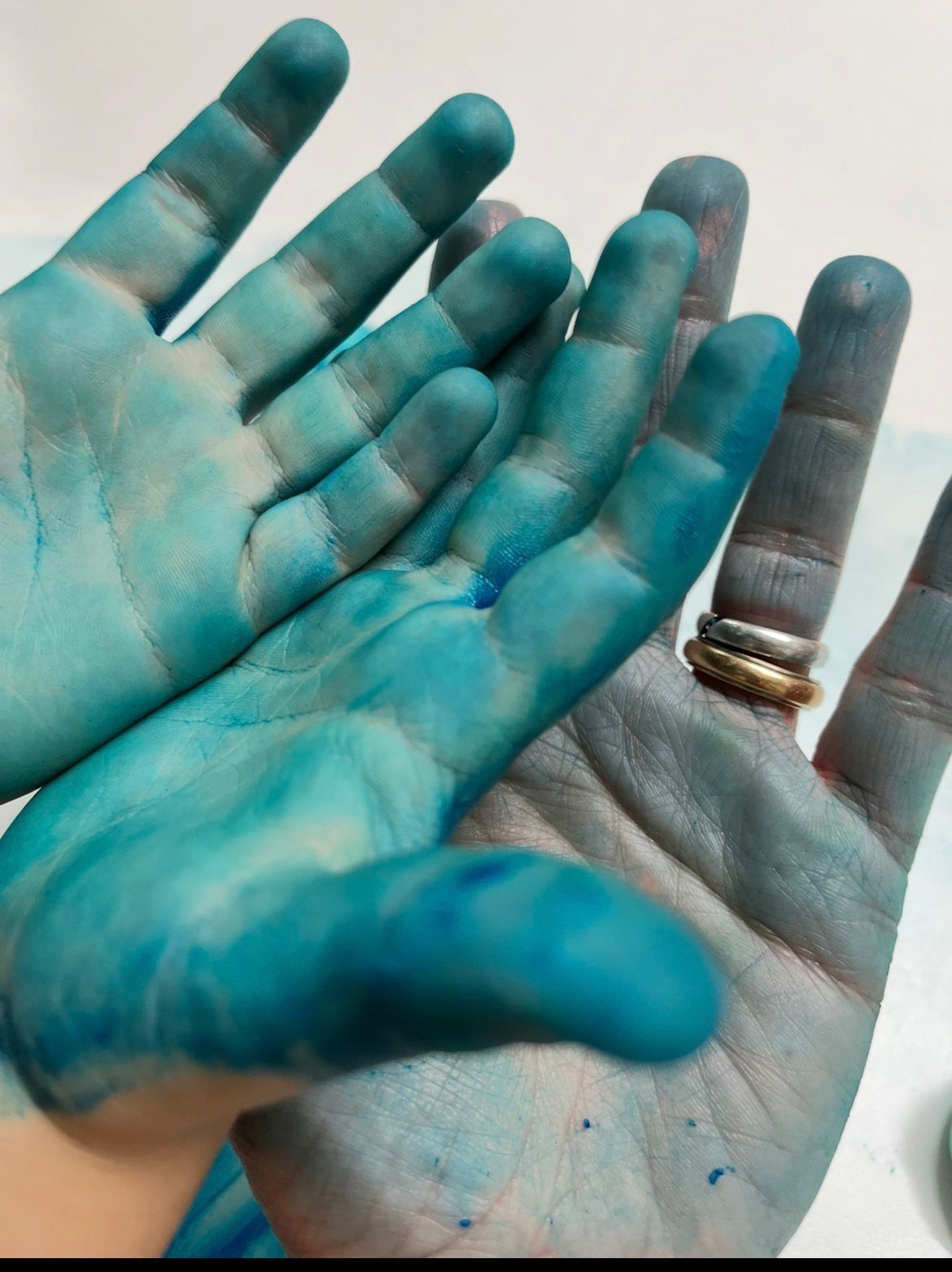 Blau bemalte Hände, die gemeinsam erhoben sind, wobei eine Hand goldene Ringe trägt, vor hellem Hintergrund