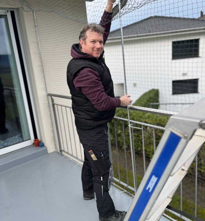 Ein Mann steht auf einem Balkon und installiert ein Katzennetz. Foto von der Stöckli Katzenschutz GmbH