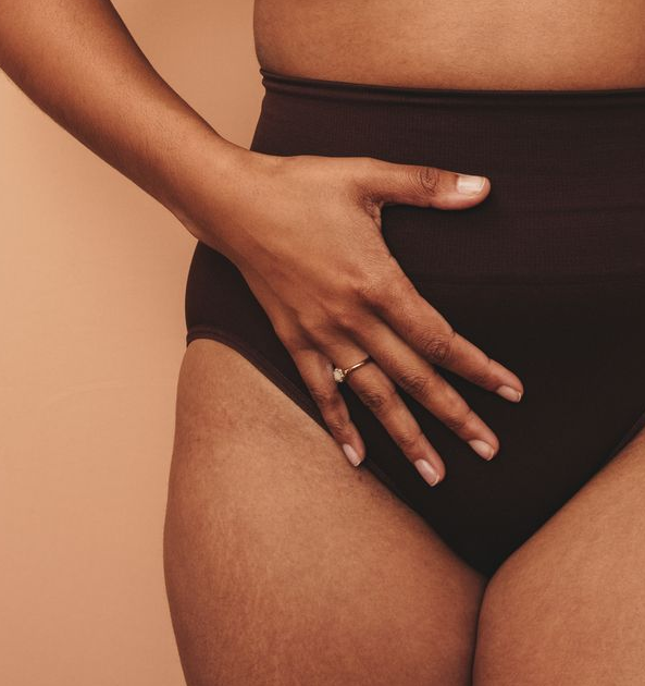 Mão sobre o abdômen, por cima de uma calcinha de cintura alta; pele morena, fundo neutro.