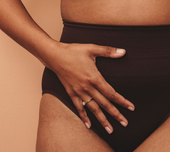Mão de pessoa sobre o abdômen, usando calcinha escura de cintura alta e com um anel no dedo. Fundo cor de pêssego.