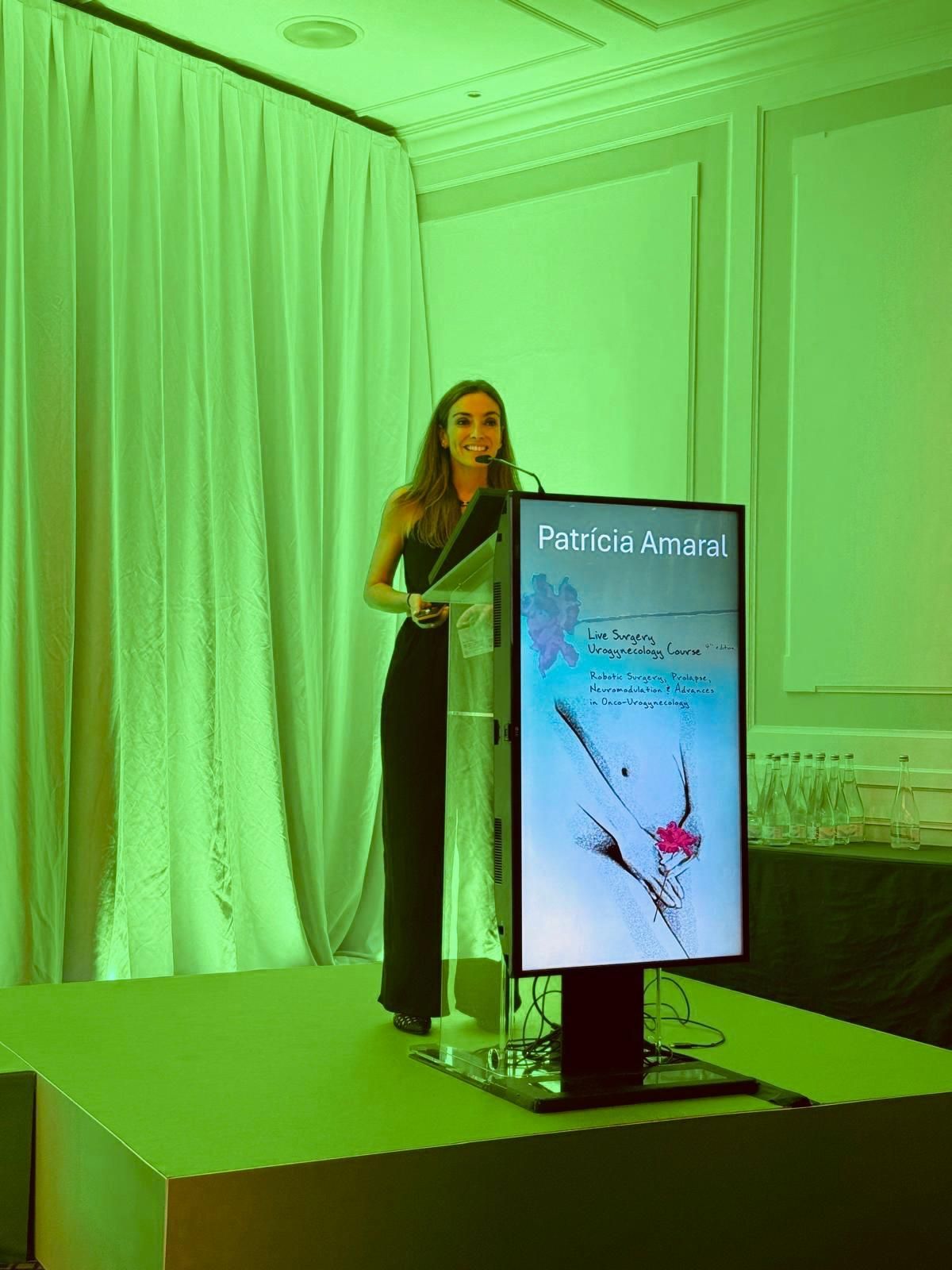 Mulher discursando em um pódio com uma tela exibindo uma ilustração médica, iluminada com luz verde.