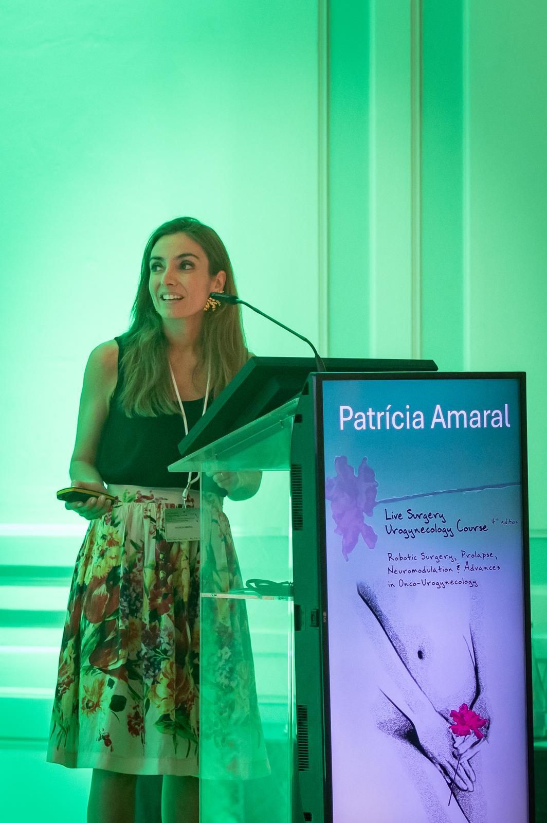 Uma mulher, Patricia Amaral, discursa de um pódio usando uma saia florida; sala com iluminação verde.