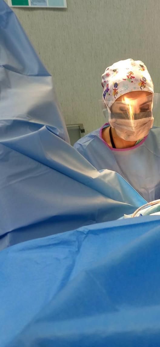 Uma pessoa vestindo roupa cirúrgica, máscara e touca está trabalhando em uma sala de cirurgia. Campos cirúrgicos azuis estão em primeiro plano.