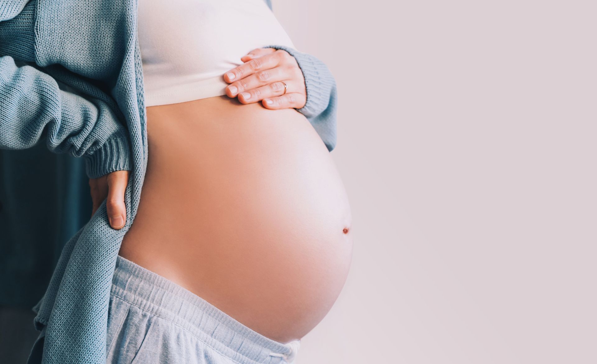 Mulher grávida, barriga à mostra. Mãos apoiadas na lateral do corpo, segurando a barriga. Suéter azul claro.