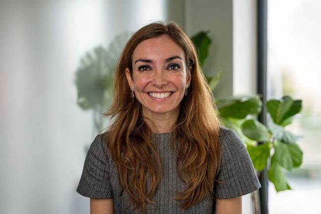 Mulher sorridente, vestindo um vestido cinza estampado, em pé em uma sala bem iluminada com plantas.