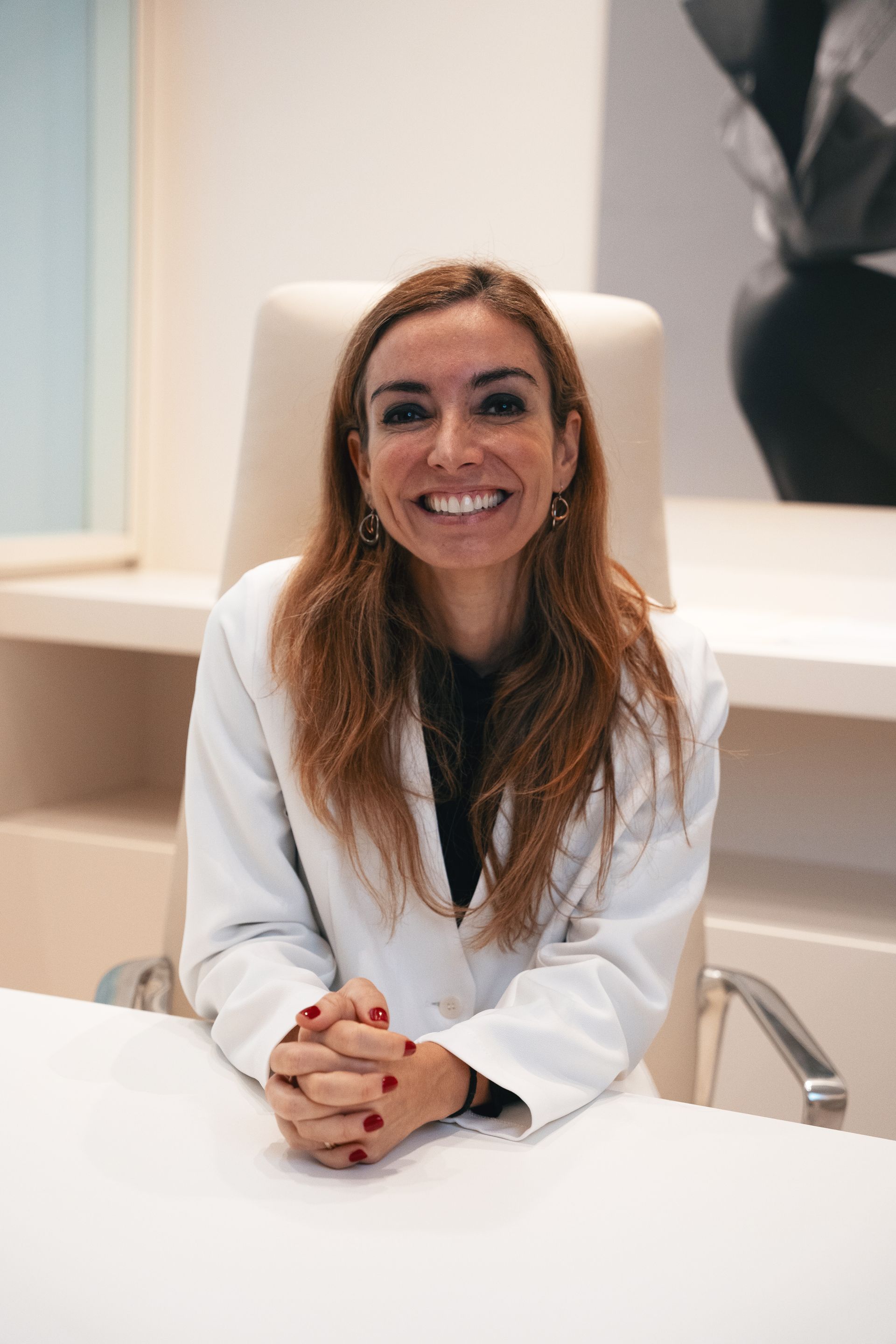 Mulher de cabelos longos e castanhos, sorrindo, vestindo uma blusa cinza, em pé em frente a um fundo de madeira.