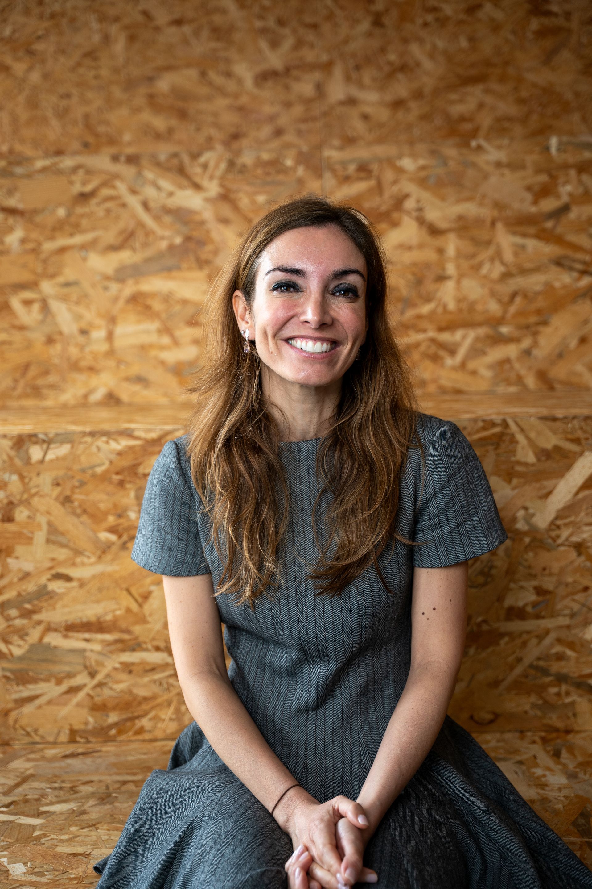 Mulher sorridente, sentada, vestindo um vestido cinza. O fundo é de madeira marrom-clara.