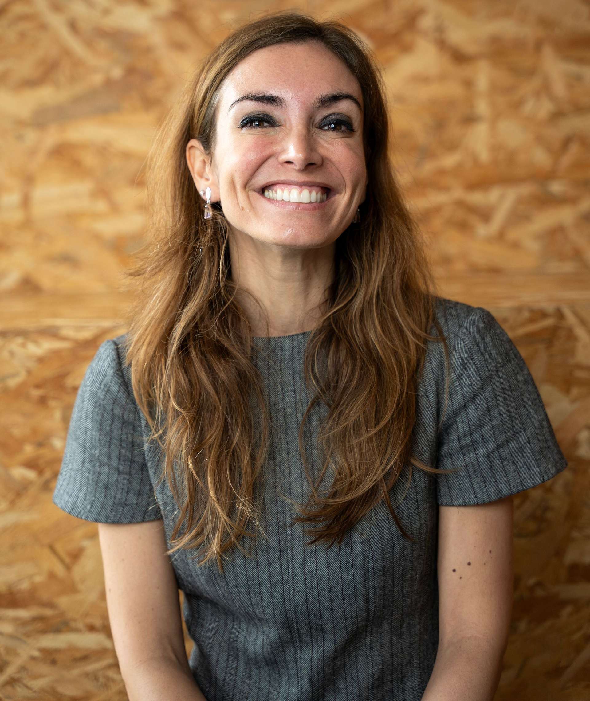 Mulher de cabelos longos e castanhos, sorrindo, vestindo uma blusa cinza, em pé em frente a um fundo de madeira.