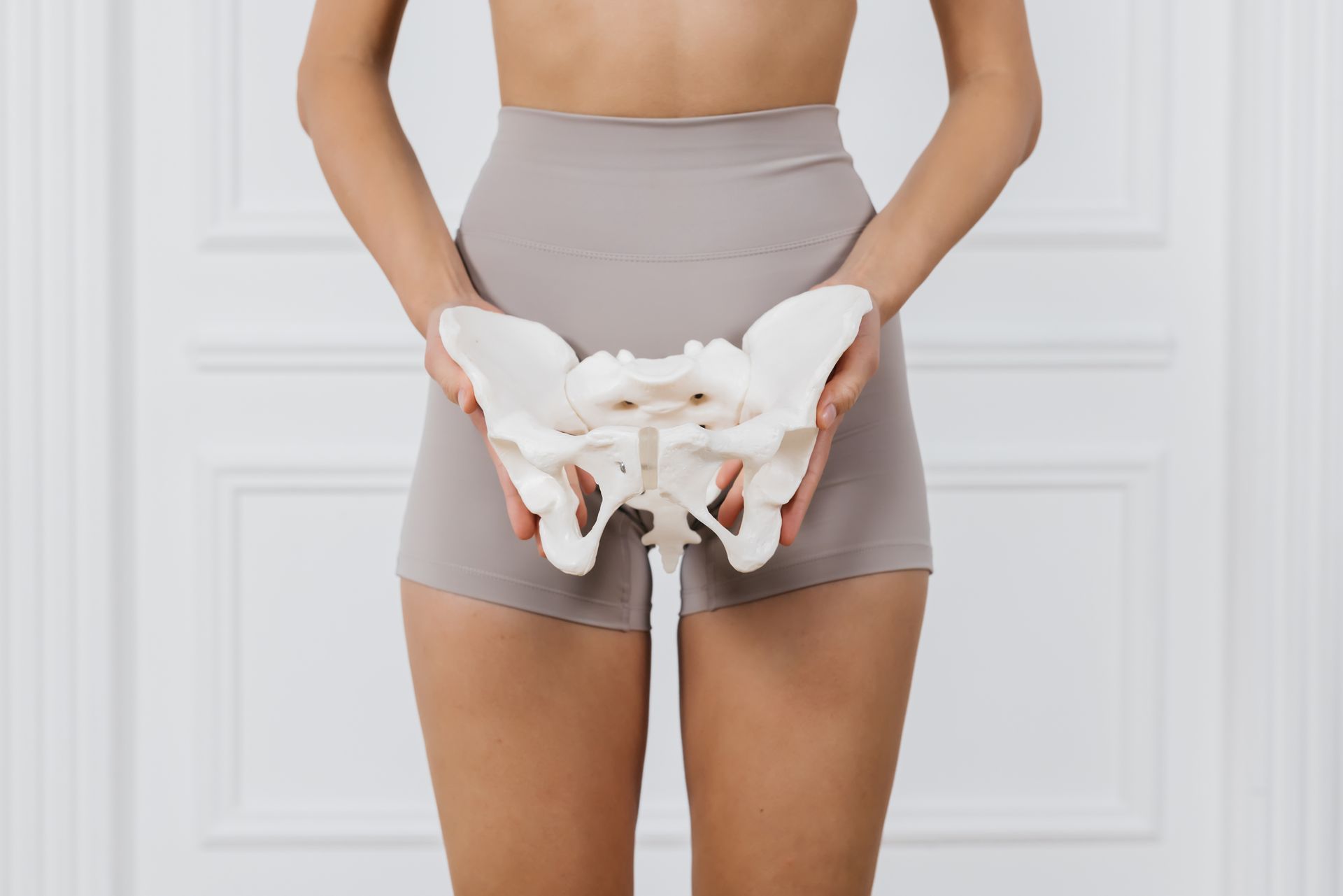 Mulher segurando uma réplica do osso pélvico em frente ao torso; vestindo shorts bege em um ambiente branco.