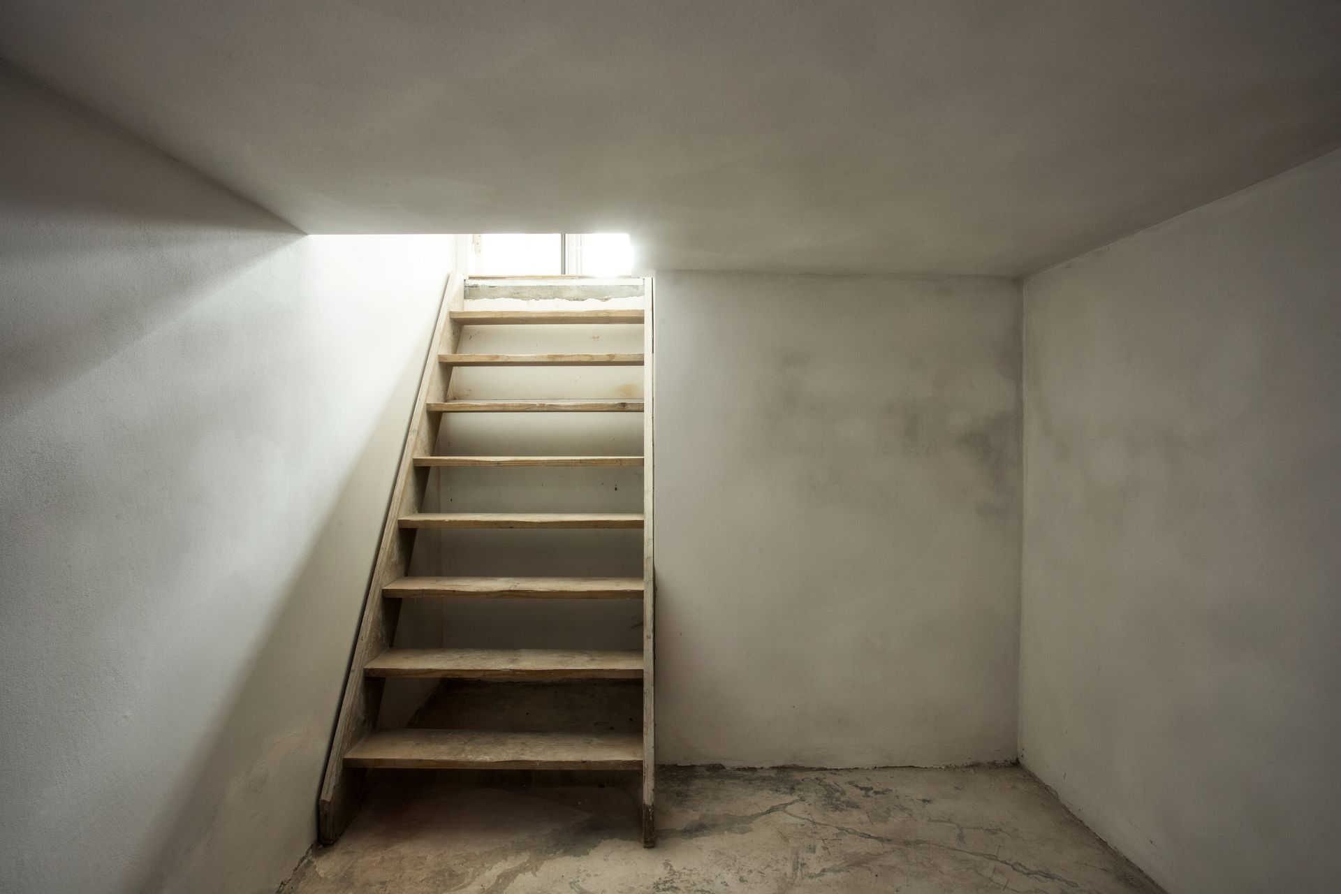 Pièce vide en sous-sol de béton avec un escalier menant à une petite ouverture lumineuse.