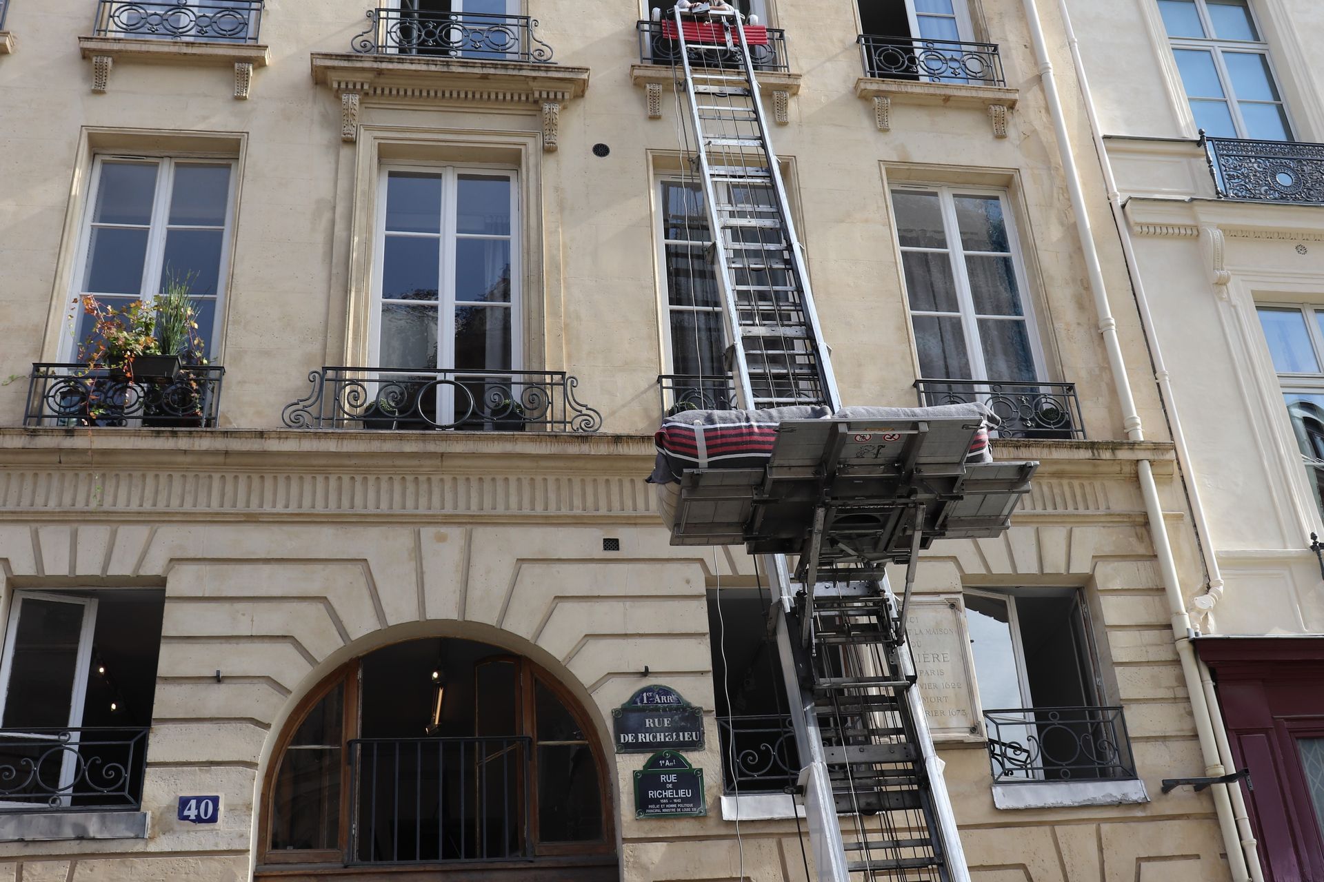 Façade d'immeuble beige dans une rue de la ville, avec un ascenseur à grande échelle desservant les étages supérieurs.