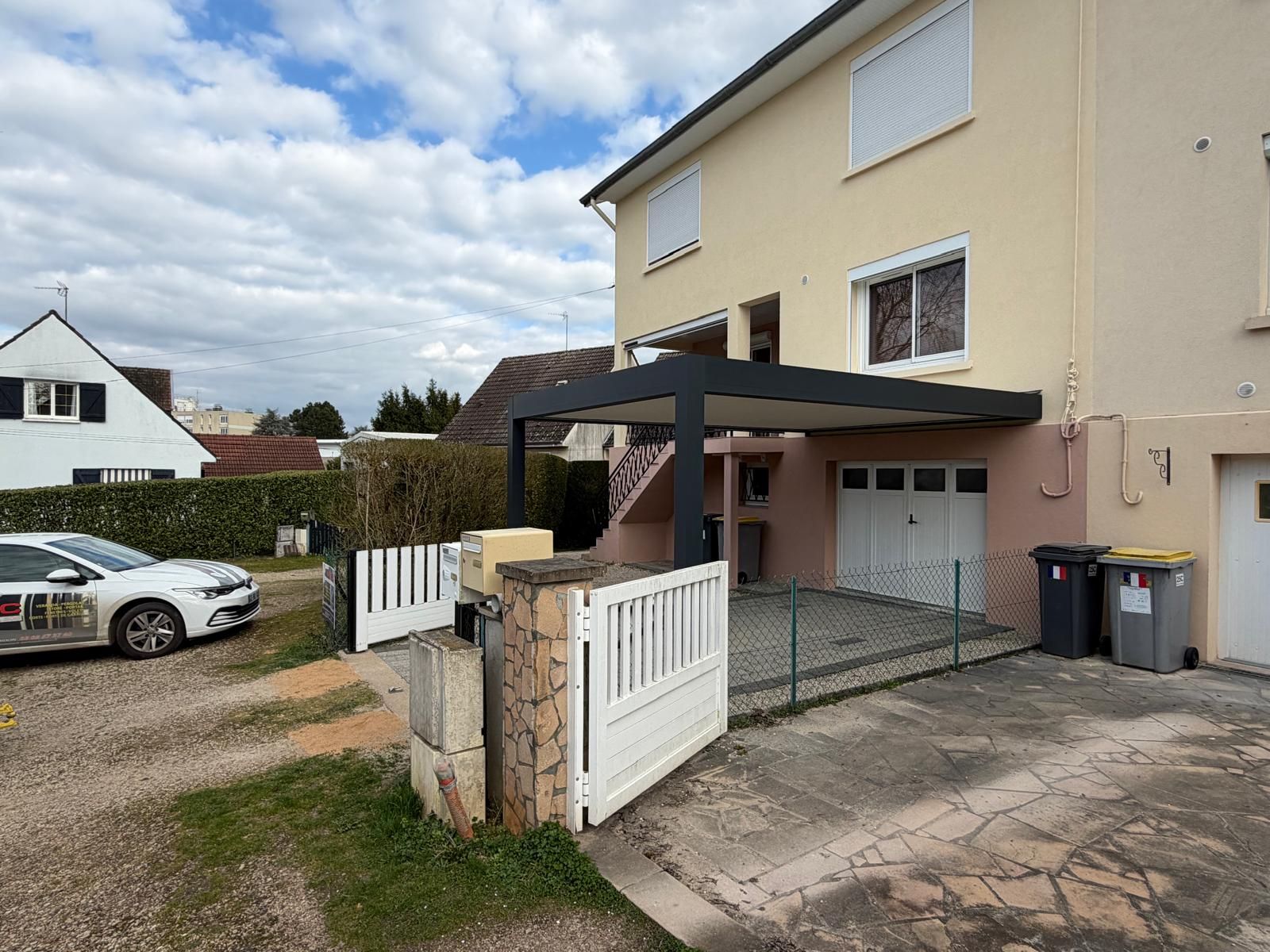 Allée résidentielle et entrée de garage jouxtant une maison beige, avec un portail blanc et une voiture garée à proximité.