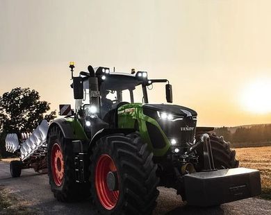 Un gran tractor verde con ruedas rojas circulando por un camino de tierra al atardecer.