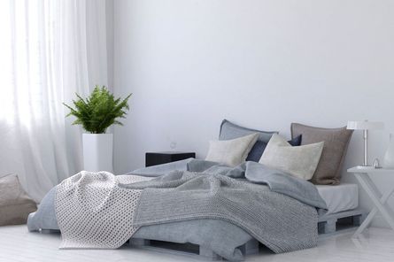 Dormitorio con paredes blancas, cama con ropa de cama y almohadas grises, planta en maceta blanca y cortinas traslúcidas.