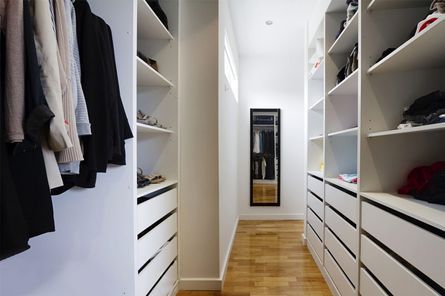 Vestidor con estantes y cajones blancos a ambos lados de un pasillo con suelo de madera; la ropa cuelga a la izquierda.