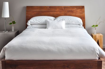 Cama de madera con ropa de cama blanca, flanqueada por una lámpara, una mesita de noche y un pequeño jarrón.