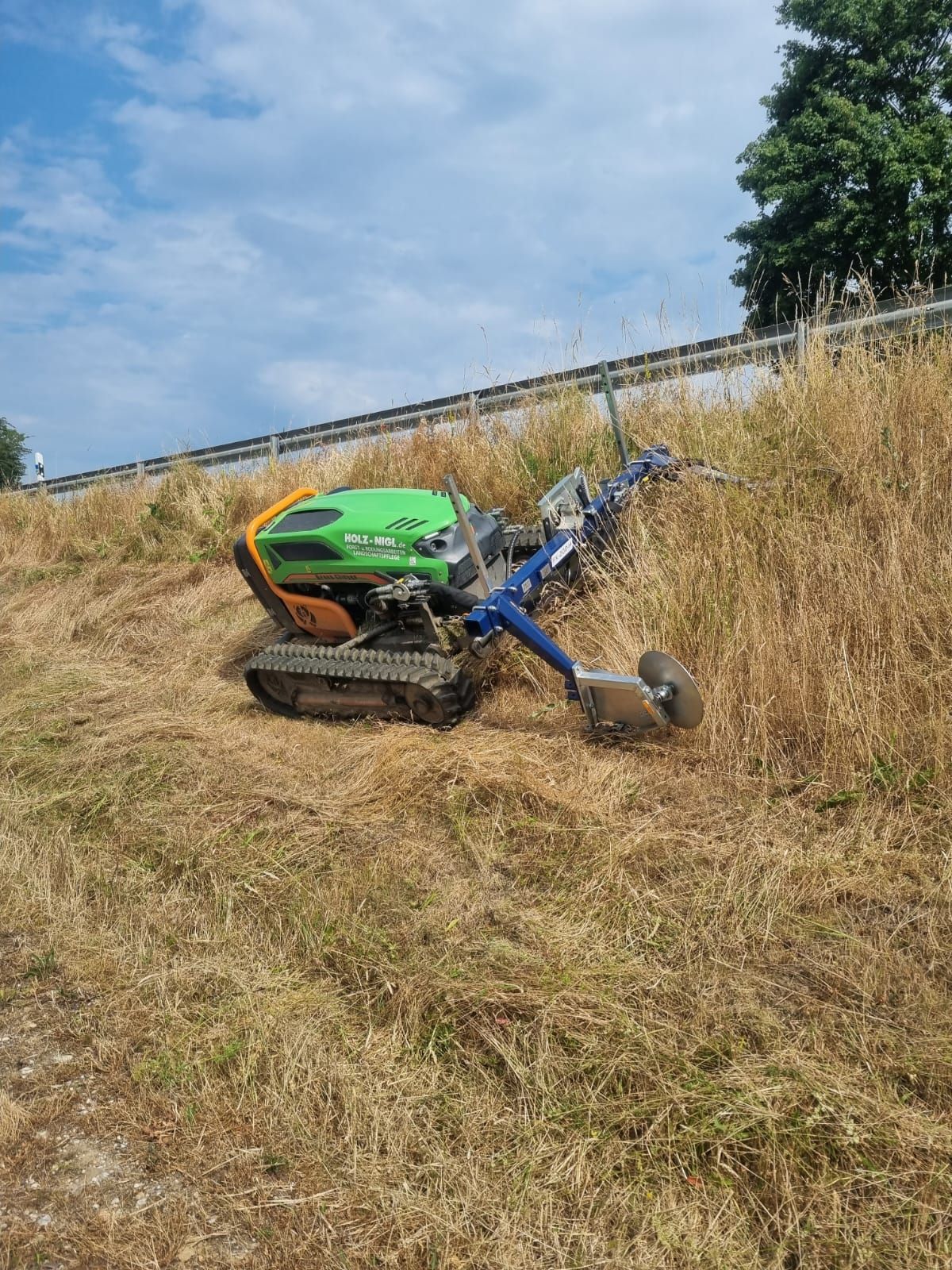 Ein Ferngesteuerter Mähroboter kürzt einen angeschrägten Hang an einer Straße