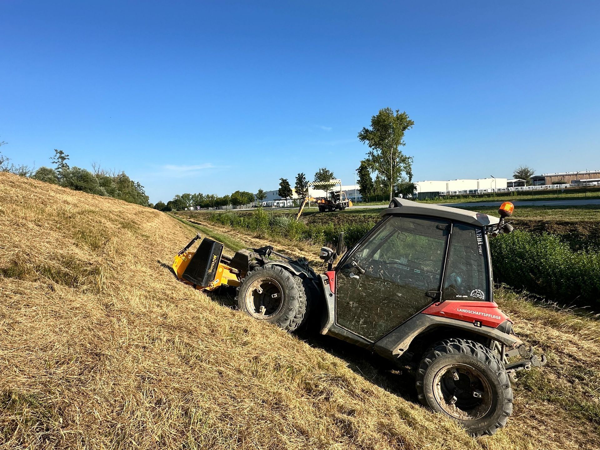 Kleines rotes Fahrzeug mäht steilen Hang an sonnigem Tag, Umgebung mit Bäumen, Straße und Gebäuden im Hintergrund.