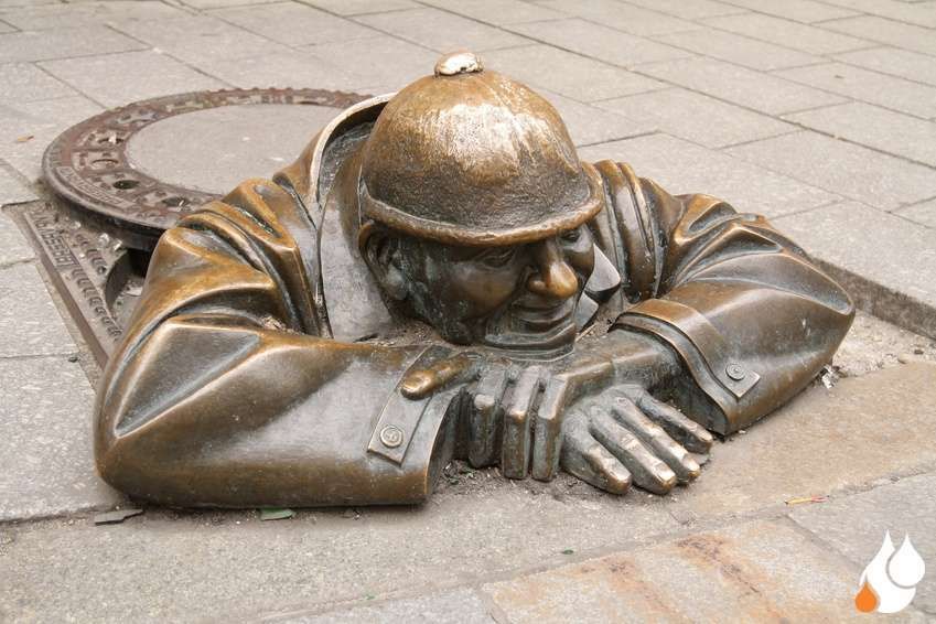 Start - Männliche Figur aus Messing guckt aus einem Gully hervor, der Gullydeckel ist beiseite geschoben