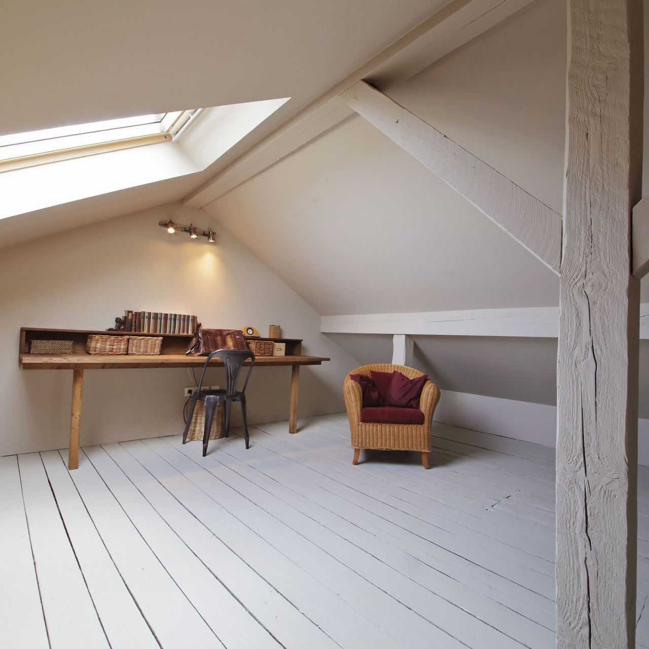 Espace de travail sous les combles avec bureau, chaise, étagères, livres et fauteuil. Sol en bois clair et murs blancs.