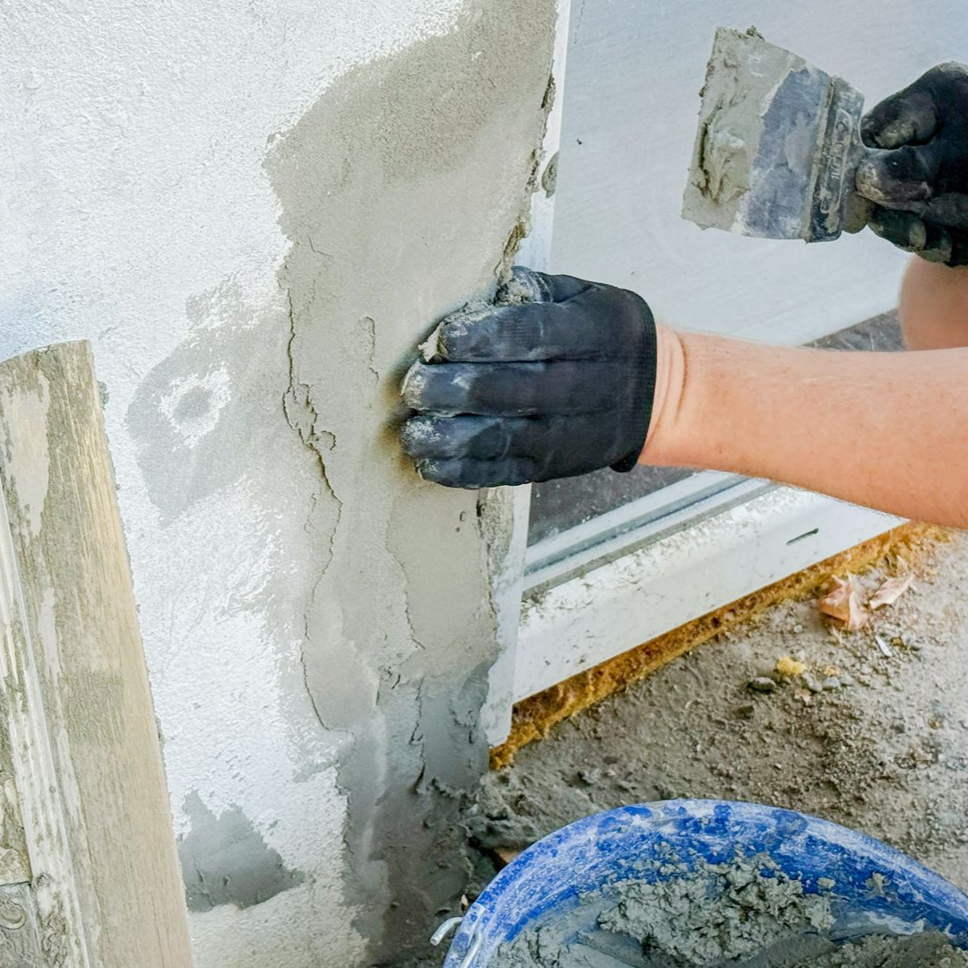 Une personne portant des gants noirs applique du plâtre sur un mur à l'aide d'une truelle près d'une fenêtre.