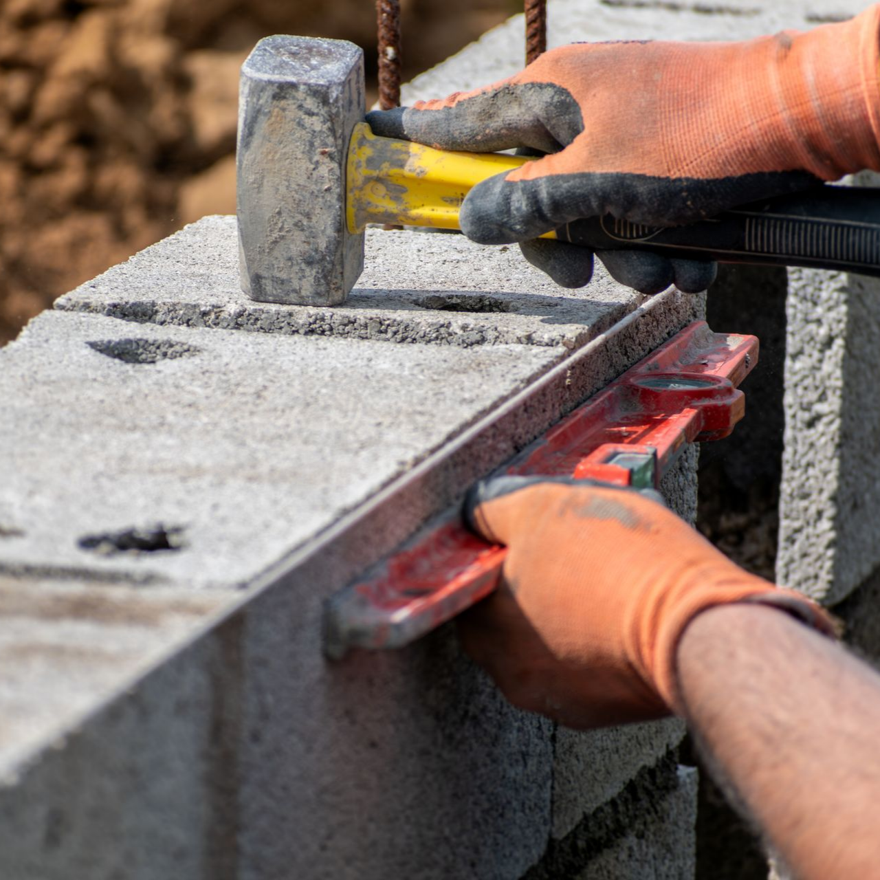 Des mains gantées utilisent un marteau et un niveau pour construire un mur en parpaings.