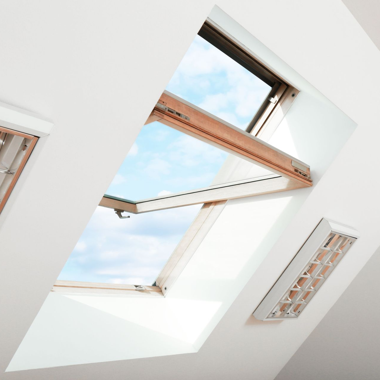 Puits de lumière ouvert sur un plafond blanc incliné, ciel bleu visible. Une grille d'aération blanche se trouve à proximité.