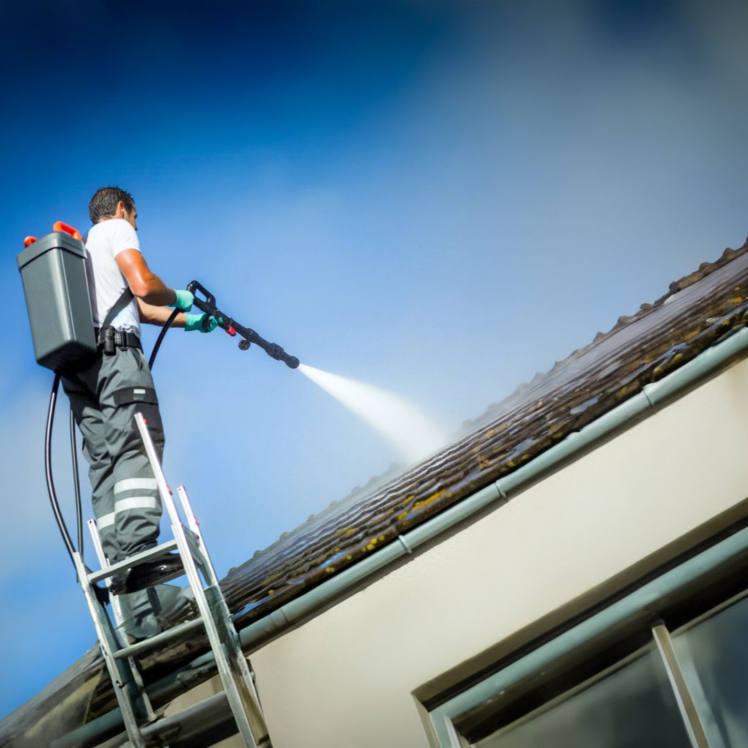 Un homme sur une échelle nettoie un toit au nettoyeur haute pression à dos, sur fond de ciel bleu éclatant.