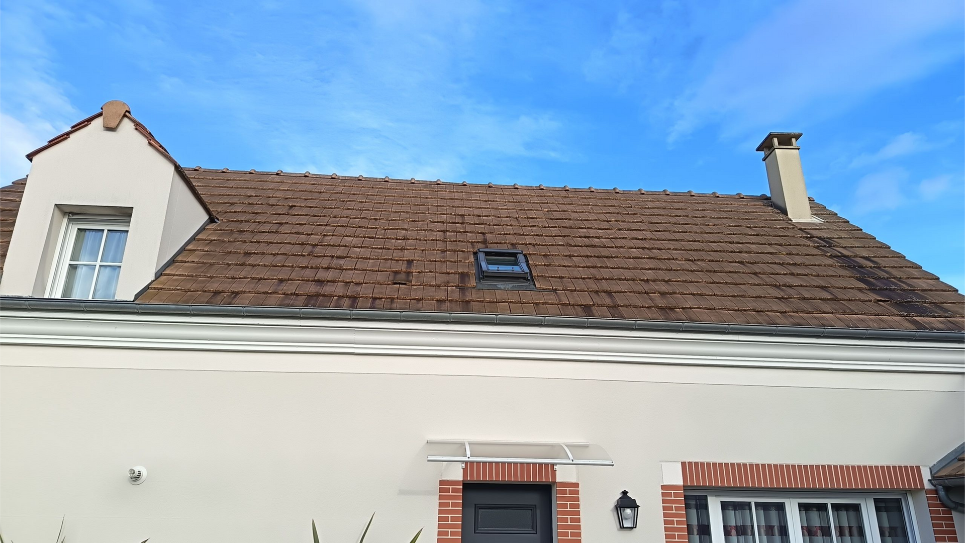 Toit en bardeaux bruns sur une maison beige se détachant sur un ciel bleu, avec une cheminée et une petite fenêtre.