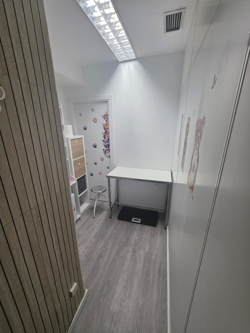 Pequeña sala de reconocimiento con báscula, mesa y mueble de almacenamiento. Una puerta con huellas dactilares da a un pasillo.