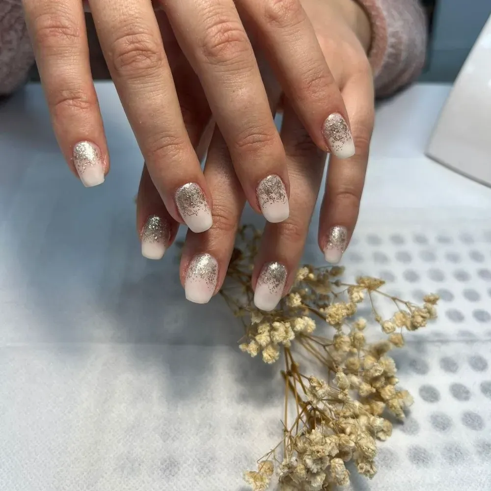 Manos con uñas de color rosa claro y puntas de brillo, apoyadas sobre una superficie blanca con flores secas.