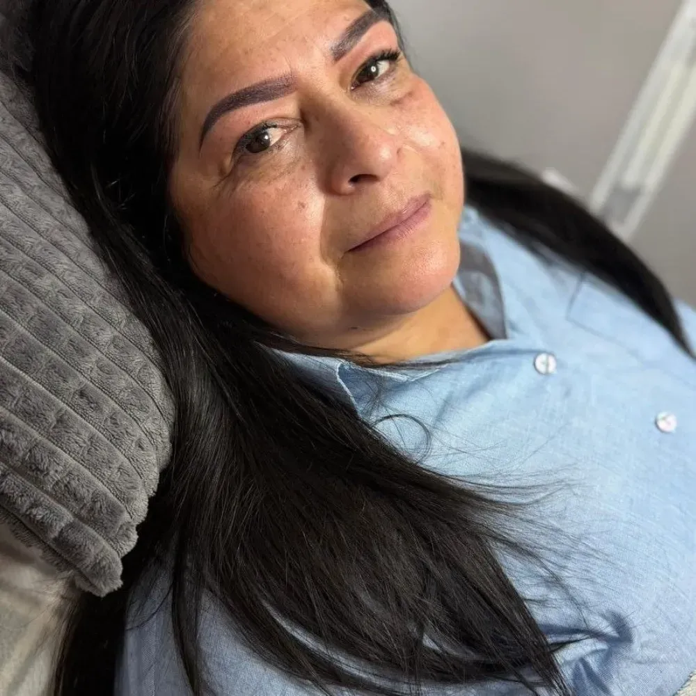 Mujer de cabello oscuro y cejas recién depiladas, vestida con una camisa azul claro abotonada, descansando sobre una almohada.