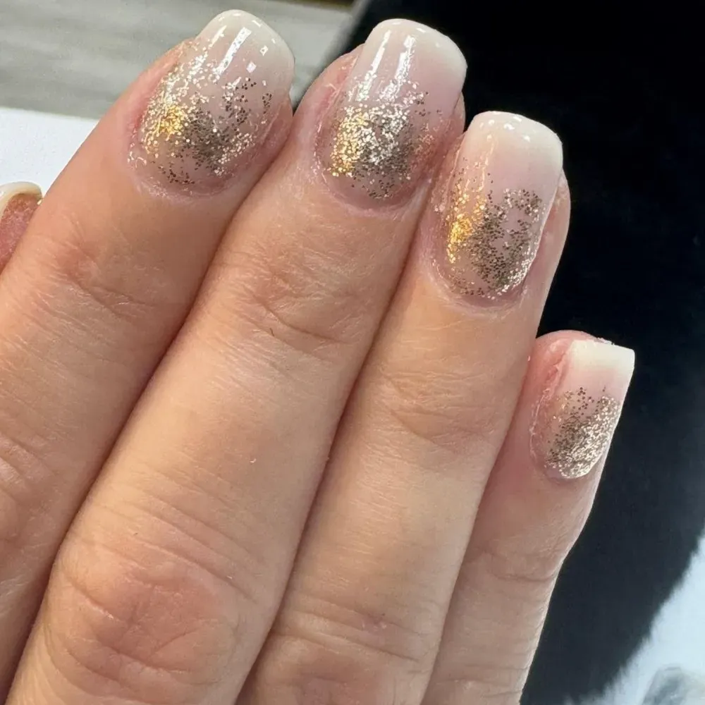 Primer plano de una mano con uñas de color rosa claro y detalles de brillo dorado en las puntas.